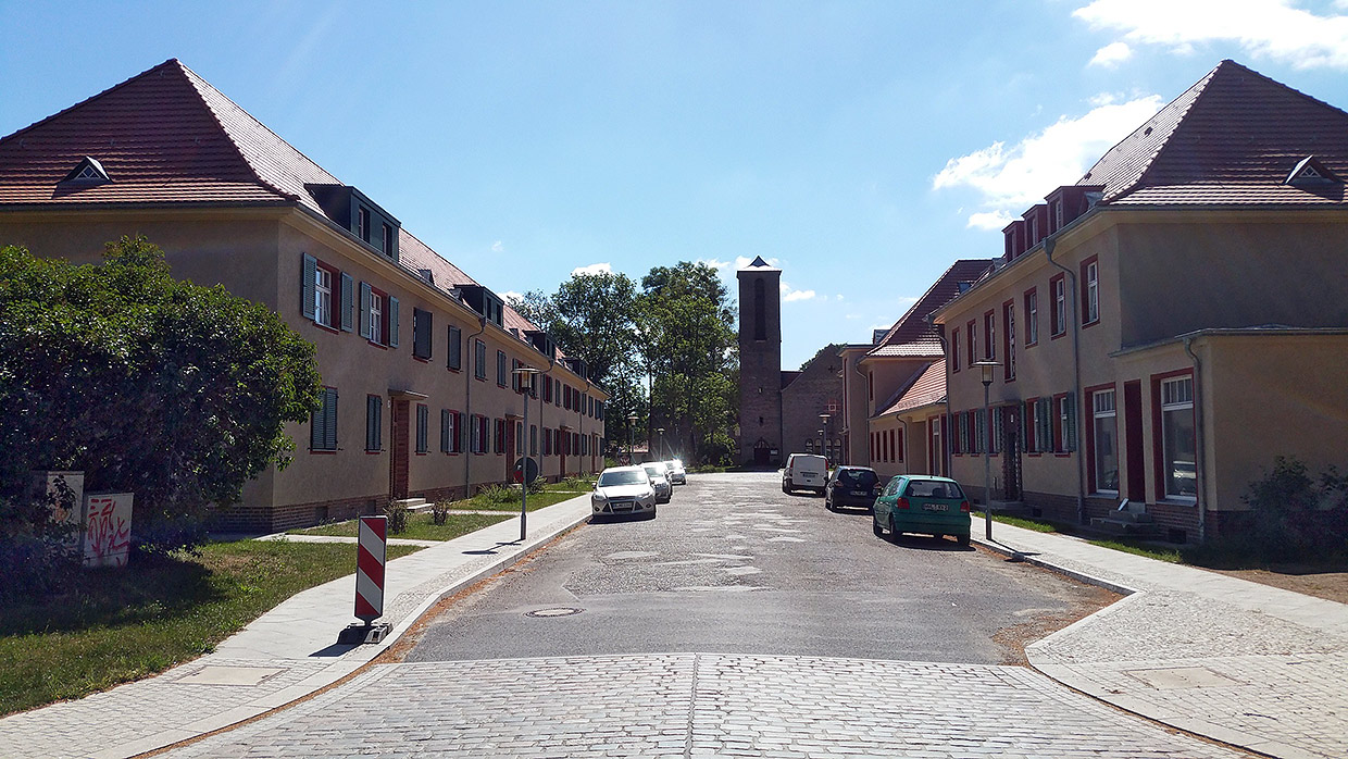 Strassenansicht am Karl-Liebknecht-Platz mit Siedlungskirche Elstal . Sanierung Eisenbahnersiedlung, Elstal/ Wustermark Strassenansicht am Karl-Liebknecht-Platz mit Siedlungskirche Elstal . Sanierung Eisenbahnersiedlung, Elstal/ Wustermark